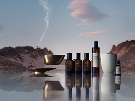 Une collection de produits parfumes Aesop pour la maison, comprenant des sprays en verre ambre, un bruleur a huile et de l’encens, disposes sur une surface reflechissante sur un paysage desertique paisible au crepuscule.